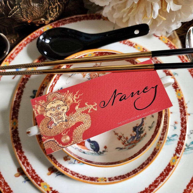 The Punctilious Mr. P's Place Card Co. "Radiant Dragon-Cinnabar" illustrated laydown place card featuring a golden-hue dragon on a chinoiserie set of china with golden chop sticks