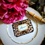 A leopard-print place card lettered with a guest's name in script, beside a pink peony and a bamboo-handled knife