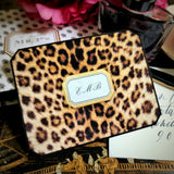 A leopard-print note card with a three-letter monogram in script within a gold-framed cartouche, propped against a crystal ink bottle, from the Punctilious Mr. P's Place Card Company