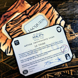 A clipped-corner card printed "MÉTIER PROVENANCE CARD" with the tiger design's story, signed in blue ink by both cofounders, over tiger-print place cards calligraphed with a guest's name in serif capitals, on a chinoiserie lacquer table