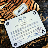 A clipped-corner card printed "MÉTIER PROVENANCE CARD" with the tiger design's story, signed in blue ink by both cofounders, over tiger-print note cards reading "Merci" in script on a chinoiserie lacquer table
