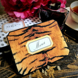 A tiger-print note card reading "Merci" in script within a gold-framed cartouche, propped beside a crystal ink bottle and pink peonies, from the Punctilious Mr. P's Place Card Company