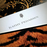 A note card personalized with a name in serif capitals beneath the anthemion motif, over tiger print