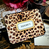 A leopard-print note card reading "MERCI" in a gold-framed cartouche, beside a crystal ink bottle and pink peonies