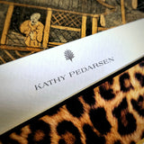 A note card personalized with a name in serif capitals beneath the anthemion motif, over leopard print