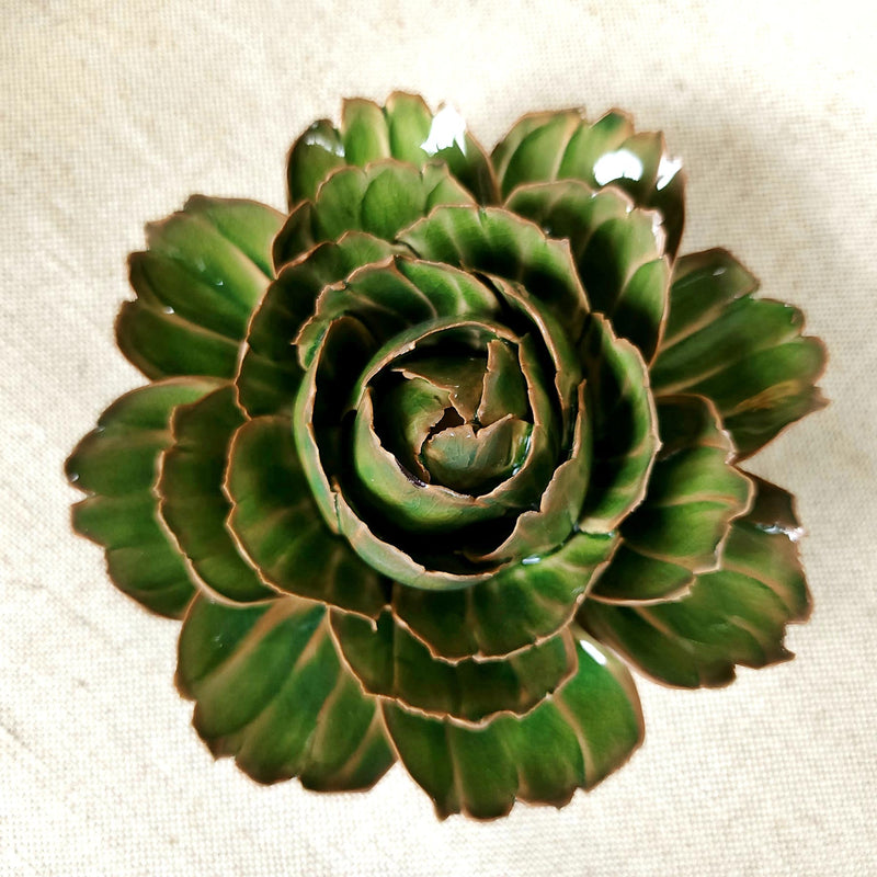 Ceramic flower resembling an olive Tipped Peony, displayed on a dinner table with green foliage and elegant tableware.