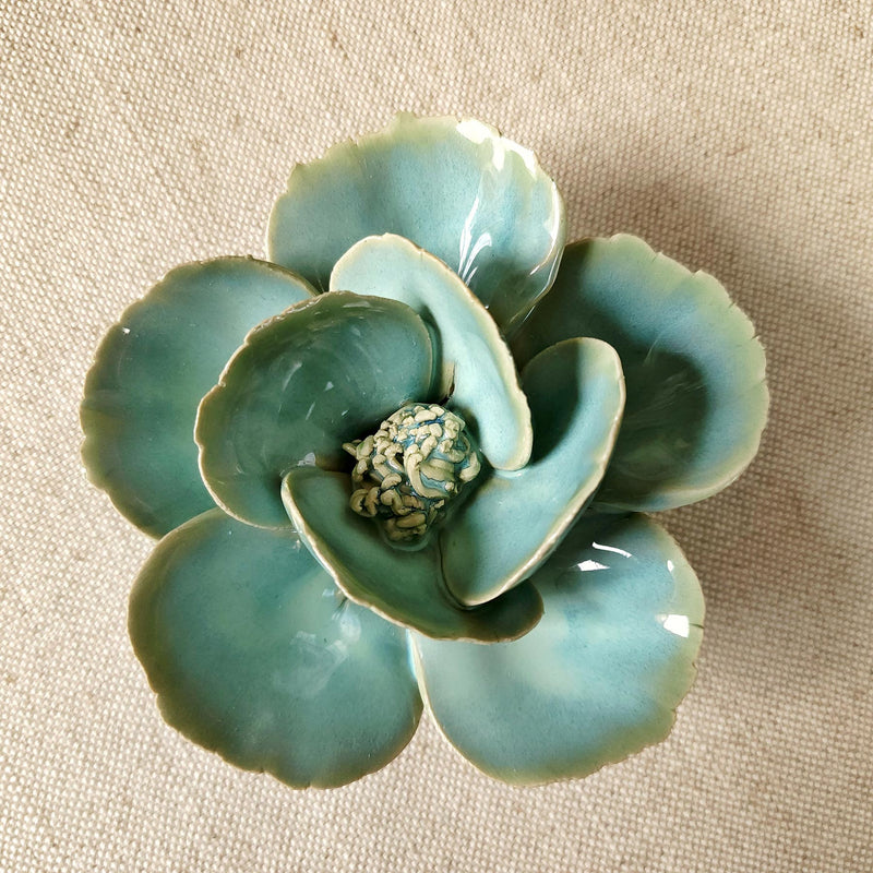 Ceramic flower resembling a celadon colored Lotus, displayed on a dinner table with green foliage and elegant tableware.