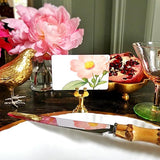 The Punctilious mr. P's Pace Card Co. "Garden Roses" custom place card on a gold place card holder with flowers and a pomegranate in the background