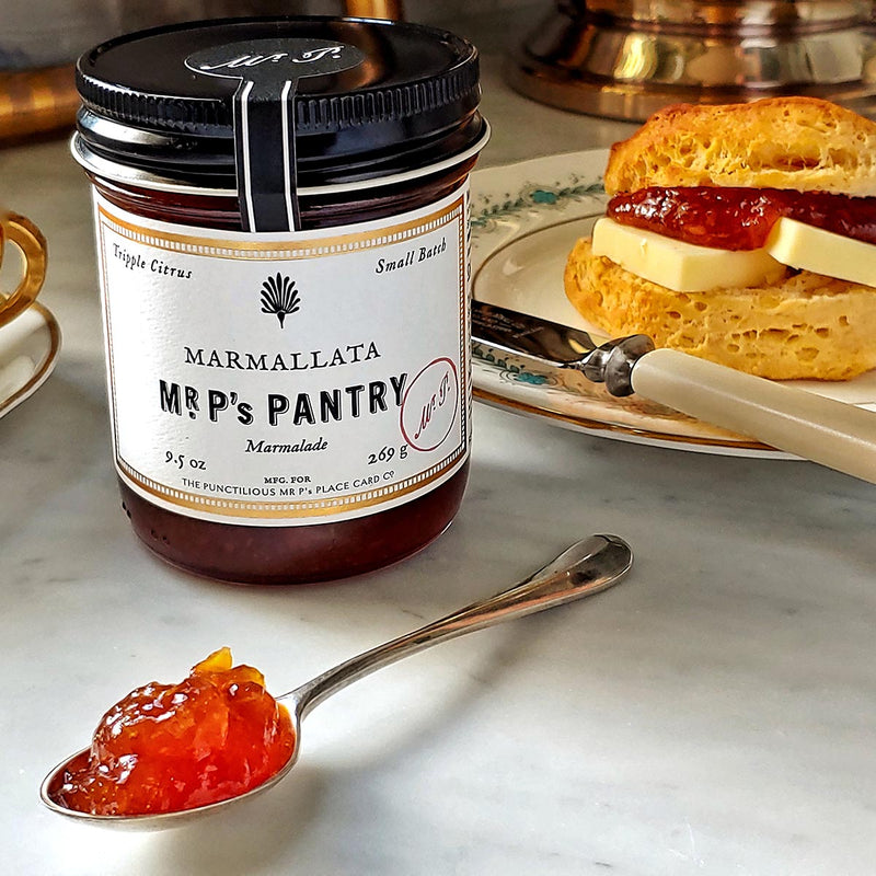 A jar of the Punctilious Mr. P's Pantry marmalade on a marble tabletop with a porcelain tea cup and saucer