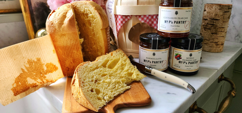 Loaf of panettone with a slice cut, jars of preserves, and a basket on a table.
