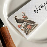 detail of The Punctilious Mr. P's Place Card Co. 'Fanciful Pheasants' custom laydown place card on top of a white dinner plate and bamboo cutlery