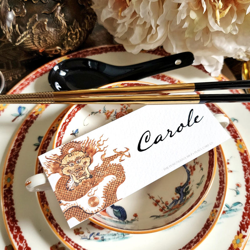 The Punctilious Mr. P's Place Card Co. "Radiant Dragon" illustrated laydown place card featuring a golden-hue dragon on a chinoiserie set of. china with golden chop sticks