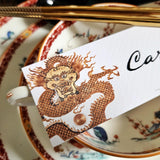 detail of The Punctilious Mr. P's Place Card Co. "Radiant Dragon" illustrated laydown place card featuring a golden-hue dragon on a chinoiserie set of. china with golden chop sticks