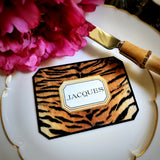 A tiger-print place card lettered with a guest's name in serif capitals, on a gold-rimmed plate with pink peonies