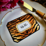A tiger-print place card with clipped corners and a two-letter monogram in a gold-framed cartouche, on a gold-rimmed plate beside a bamboo-handled knife, from the Punctilious Mr. P's Place Card Company