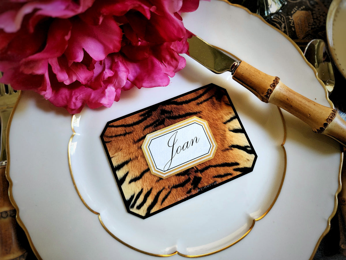 A tiger-print place card with clipped corners, a guest's name calligraphed in script within its gold-framed cartouche, on a gold-rimmed plate beside a bamboo-handled knife and a magenta peony