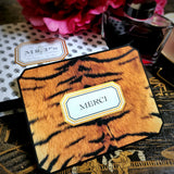 A tiger-print note card reading "MERCI" in serif capitals within a gold-framed cartouche, against the anthemion-print folio