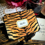 A tiger-print note card with a three-letter monogram in serif capitals, beside a fountain pen