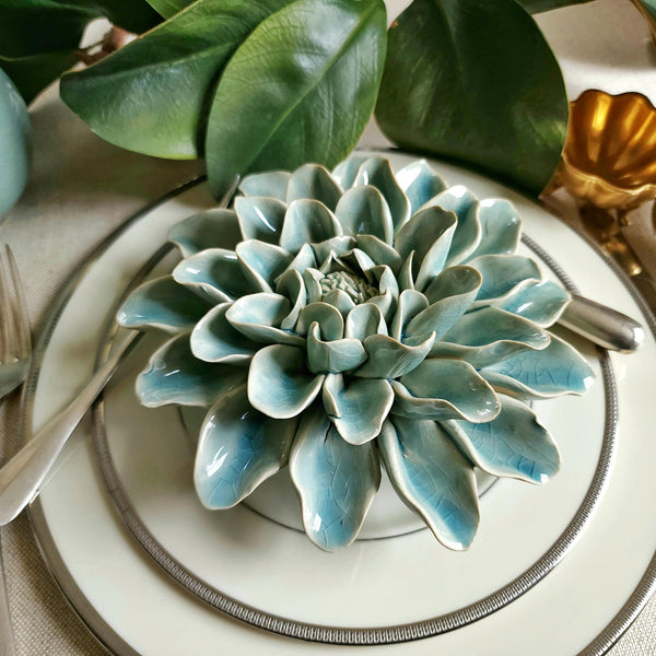 Ceramic flower resembling a celadon Dinner Plate Dahlia, displayed on a dinner table with green foliage and elegant tableware.
