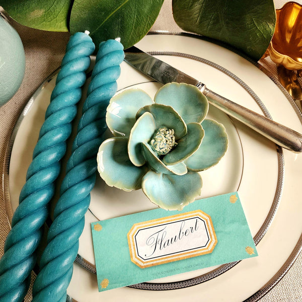 Ceramic flower resembling a celadon colored Lotus, displayed on a dinner table with green foliage and elegant tableware with Jardin colored Envoy laydown place card and matching rope candles