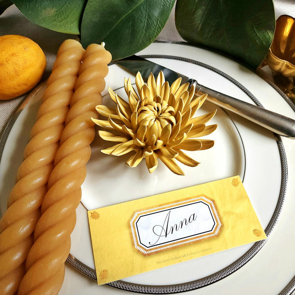 Ceramic flower resembling a yellow colored Mum, displayed on a dinner table with green foliage and elegant tableware with soleil colored Envoy laydown place card
