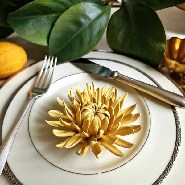 Ceramic flower resembling a yellow colored Mum, displayed on a dinner table with green foliage and elegant tableware.