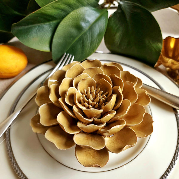 Ceramic flower resembling an caramel colored Peony, displayed on a dinner table with green foliage and elegant tableware.