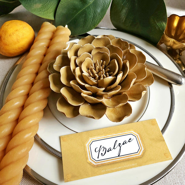 Ceramic flower resembling an olive Tipped Peony, displayed on a dinner table with green foliage and elegant tableware with soleil colored Envoy laydown place card with matching rope candle