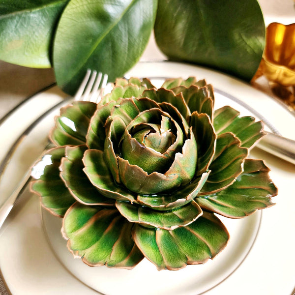Ceramic flower resembling an olive Tipped Peony, displayed on a dinner table with green foliage and elegant tableware.