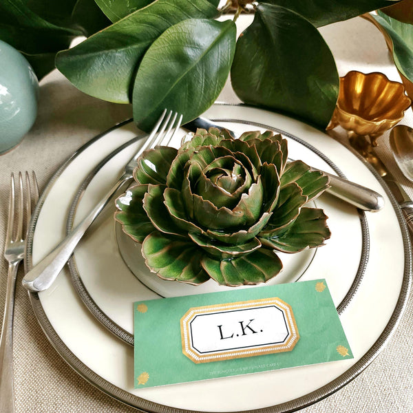 Ceramic flower resembling an olive Tipped Peony, displayed on a dinner table with green foliage and elegant tableware with Jardin colored Envoy laydown place card