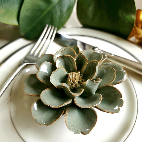 Ceramic flower resembling a sage colored Tipped Rose, displayed on a dinner table with green foliage and elegant tableware.