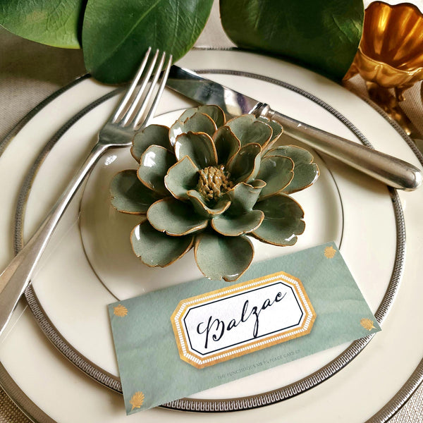 Ceramic flower resembling a sage colored Tipped Rose, displayed on a dinner table with green foliage and elegant tableware with Jardin colored Envoy laydown place card