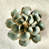 detail of the Ceramic flower resembling a sage colored Tipped Rose, displayed on a dinner table with green foliage and elegant tableware.