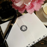 The Punctilious Mr. P x Sarah V Battle fine note card set featuring the packaging of the envelopes with sun face sealing wafer, on chinoiserie table with Mr. P's I conic black and white anthemion print. montblanc fountain pen resting on top.