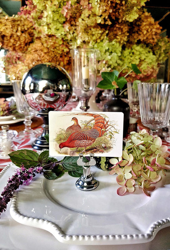 A vintage-style place card featuring an illustration of a pheasant, displayed on a silver pedestal stand on a white plate in a decorated dining table setting.