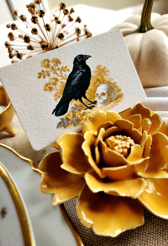 Nevermore Raven place card featuring black ravens, skulls, and green and golden botanicals, styled with a marigold ceramic blossom on an autumn table.