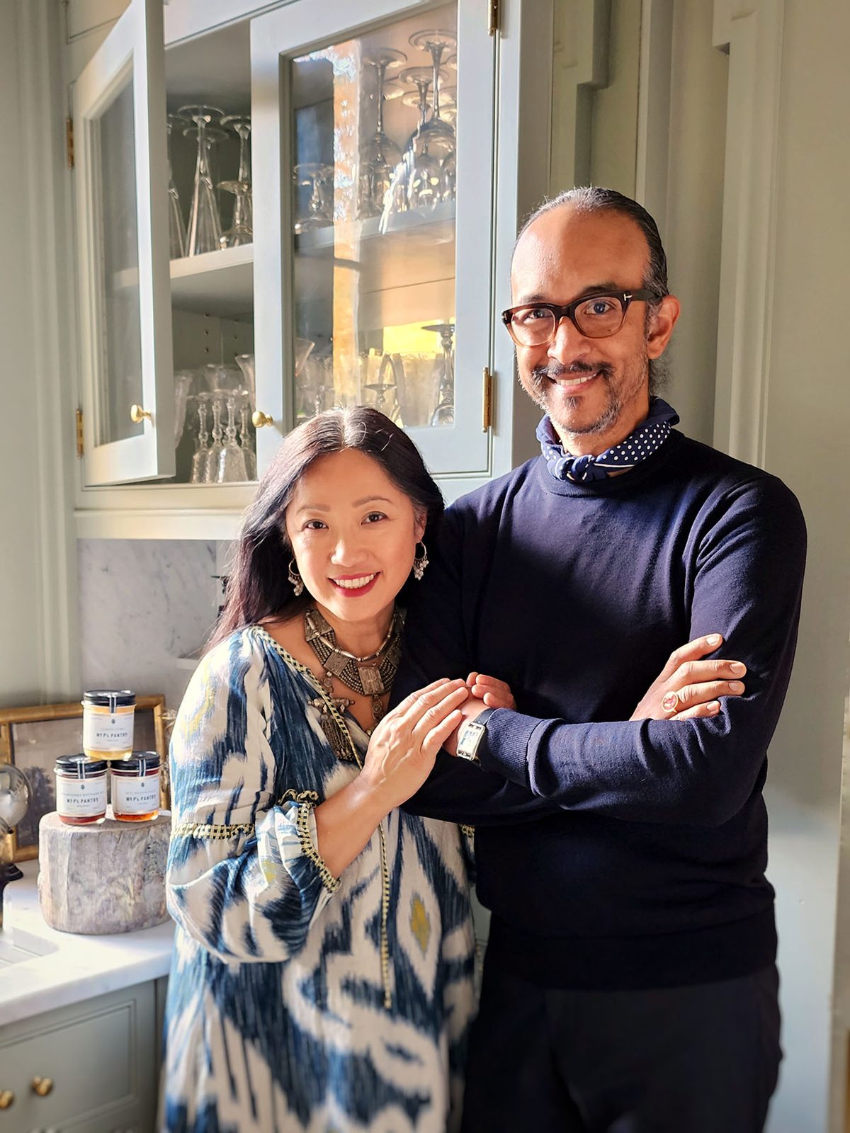 Karen Suen-Cooper and Martin Cooper, co-founders of the Punctilious Mr. P's Place Card Company, arm in arm before glass-fronted cabinets of stemware, with jars of Mr. P's Pantry on the marble counter beside them