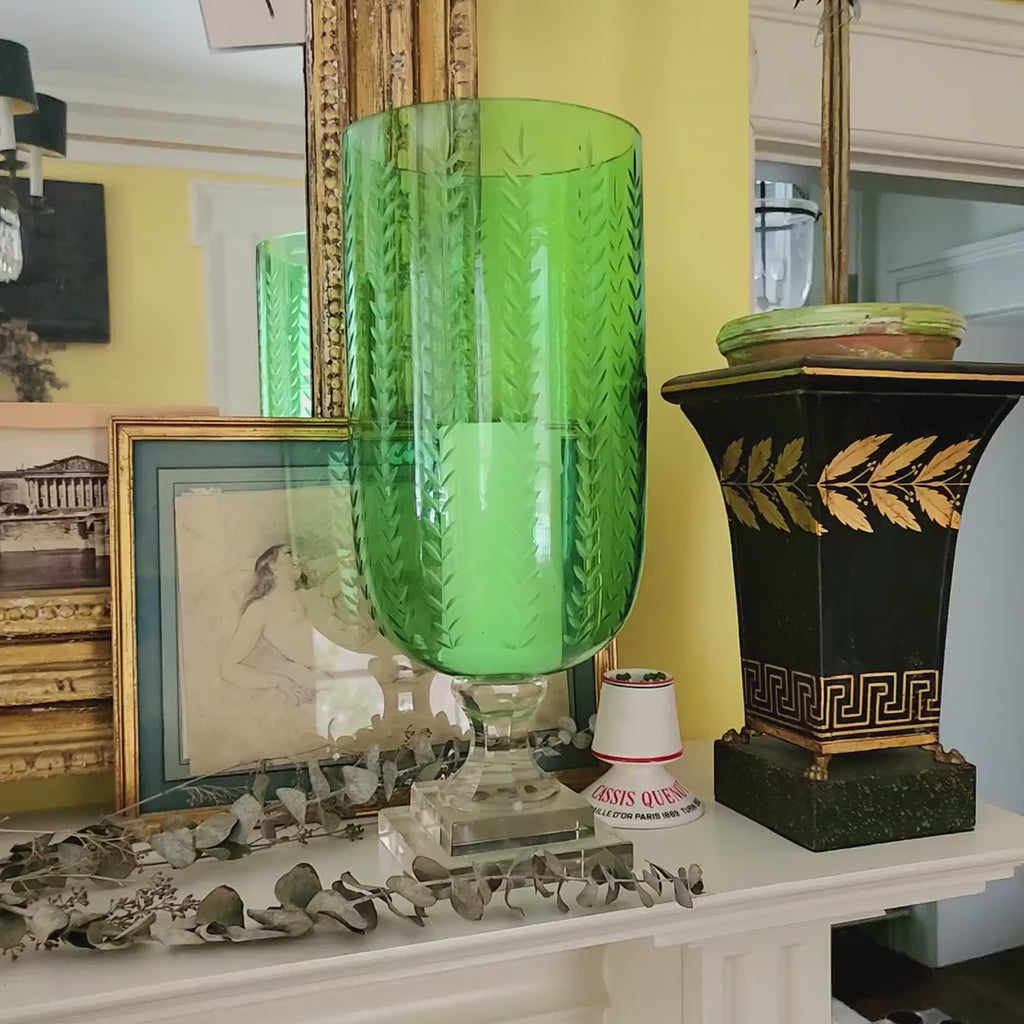 Green glass hurricane vase on a decorative shelf with framed pictures and a chandelier in the background.