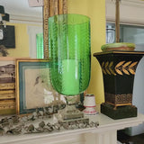 Green glass hurricane vase on a decorative shelf with framed pictures and a chandelier in the background.