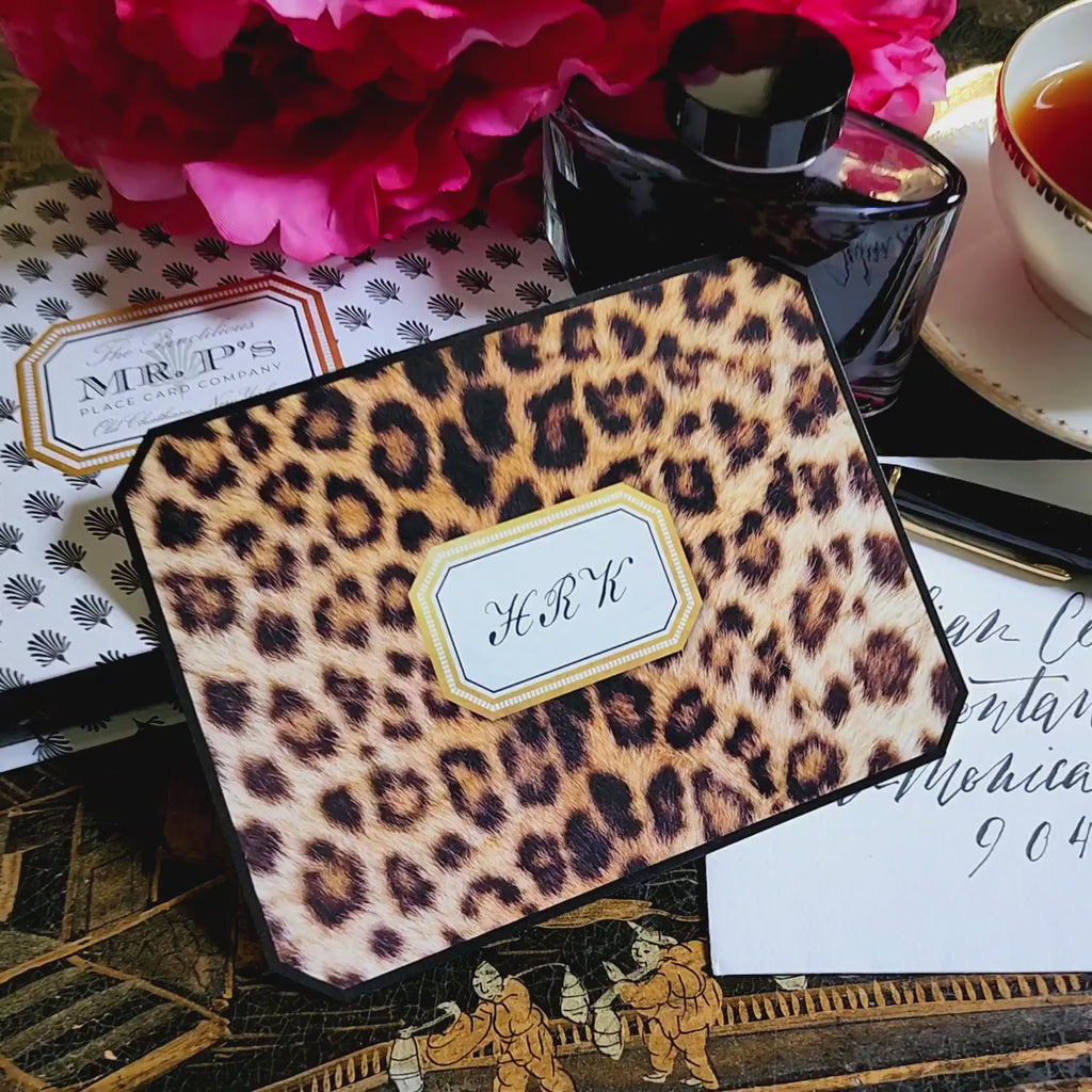 A leopard-print note card with a two- to three-letter monogram in serif capitals within its gold-framed cartouche, resting on the anthemion-print folio beside a crystal ink bottle and a calligraphed envelope