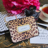 A leopard-print note card reading "MERCI" within its gold-framed cartouche, resting on the anthemion-print folio beside a crystal ink bottle, a fountain pen and a calligraphed envelope