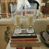 Decorative objects including a large glass greek key hurricane vase, framed artwork, and a black urn on a yellow wall background.