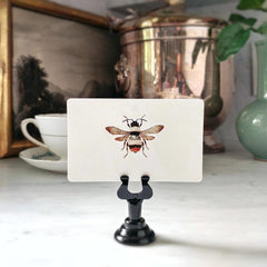 An upright Spring Bees place card illustrated with a banded bumblebee, in Mr. P's black place card holder before a teacup and a landscape painting, from the Punctilious Mr. P's Place Card Company
