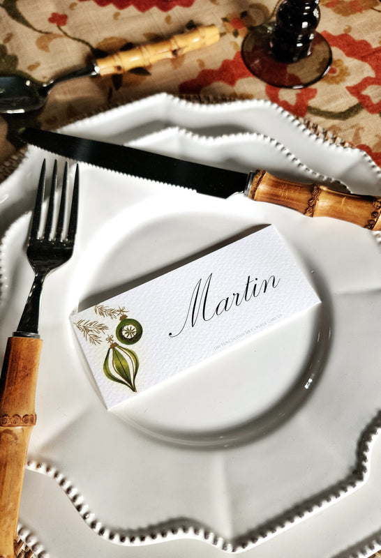 A personalized place card for 'Martin' with mid-centuray bauble illustrations from The Punctilious Mr. P's Place Card Co., folded over a white ceramic plate on a styled table setting with bamboo-handled cutlery.