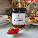 A jar of the Punctilious Mr. P's hot pepper jelly on a marble tabletop with a porcelain tea cup and saucer, and cracker in the background