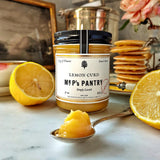 A jar of the Punctilious Mr. P's lemon curd on a marble tabletop with a wedge of lemon and a stack of silver dollar pancakes