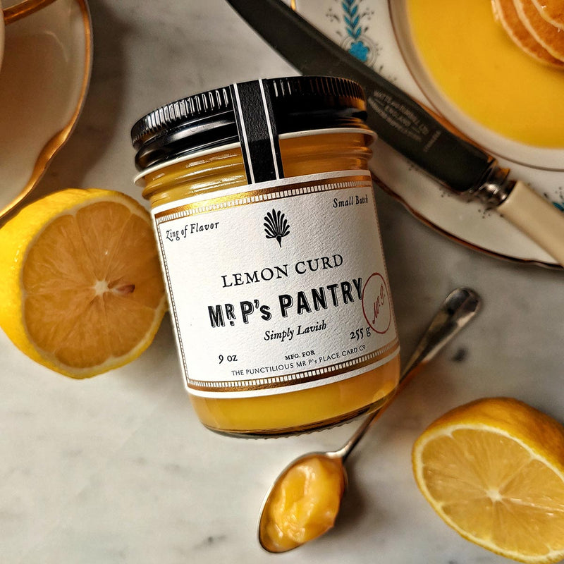 A jar of the Punctilious Mr. P's lemon curd on a marble tabletop with a wedge of lemon and a stack of silver dollar pancakes