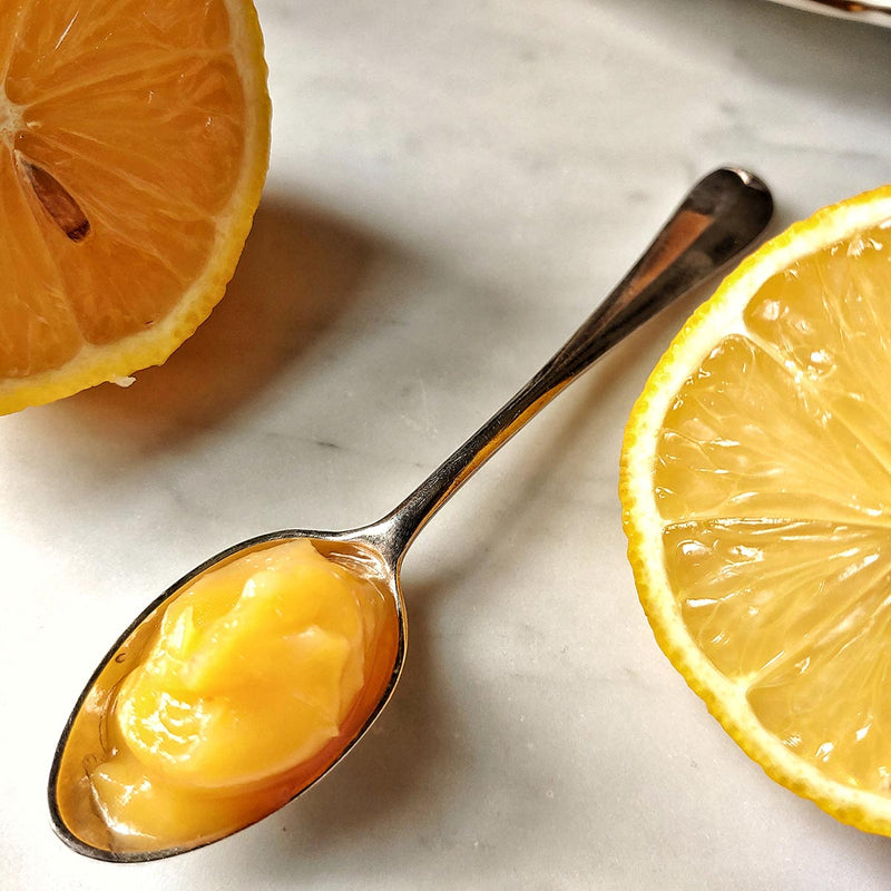 A jar of the Punctilious Mr. P's lemon curd on a marble tabletop with a dollop of lemon curd on a spoon
