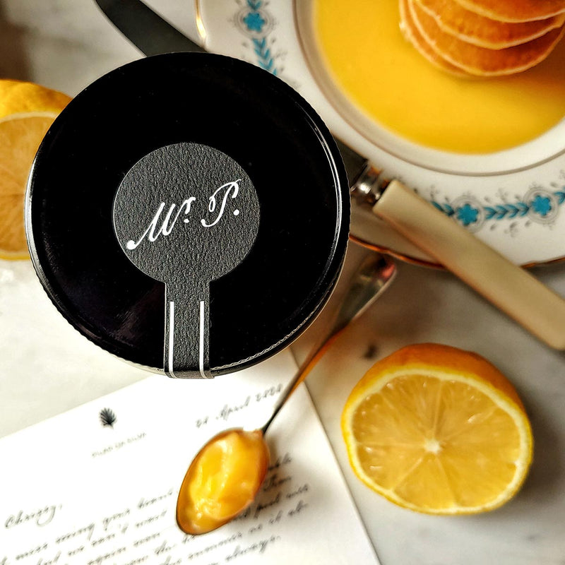jar top showing the mr. p logo of the Punctilious Mr. P's lemon curd on a marble tabletop with a wedge of lemon and a stack of silver dollar pancakes