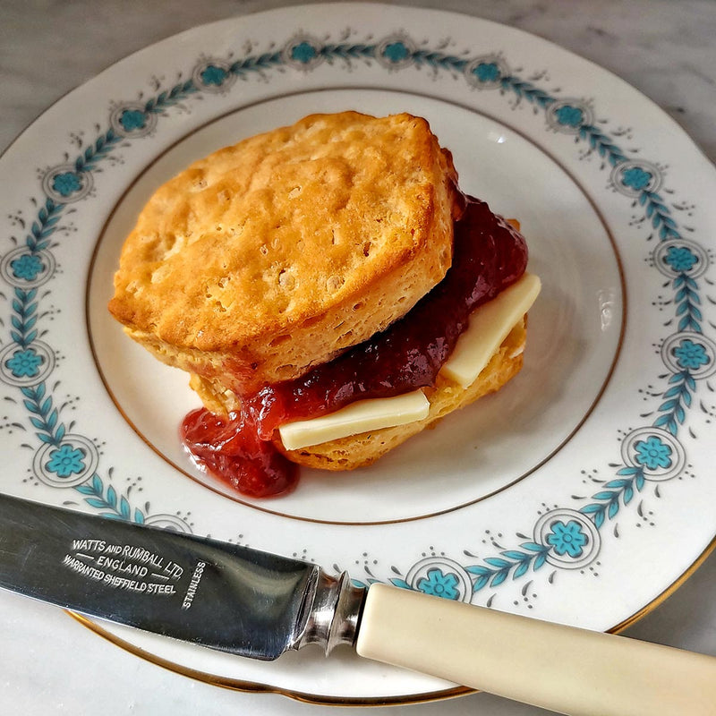 A jar of the Punctilious Mr. P's strawberry-rhubard jam on a marble tabletop with a porcelain tea cup and saucer, and biscuit in the background